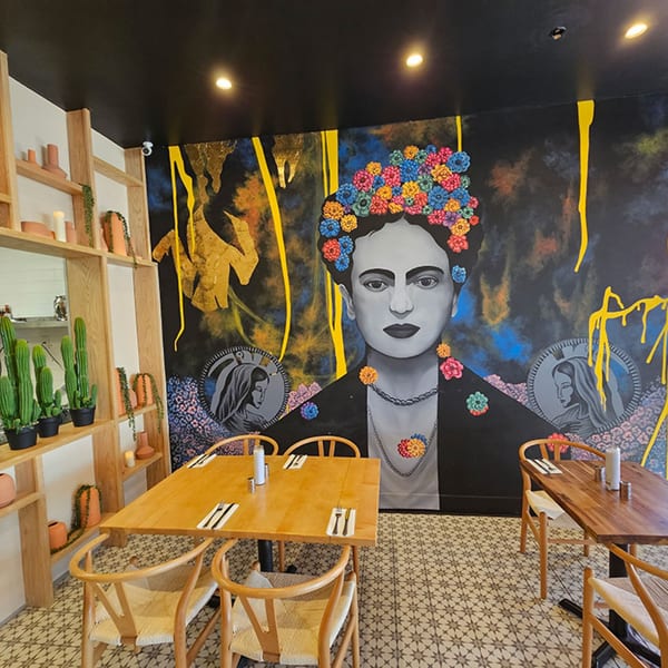 Okay Maguey dining room with a large Frida Kahlo mural, wooden tables, and shelves with cacti.