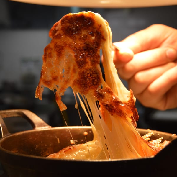 A hand pulls a spoonful of gooey, melted, browned cheese from a dark cast iron pan.