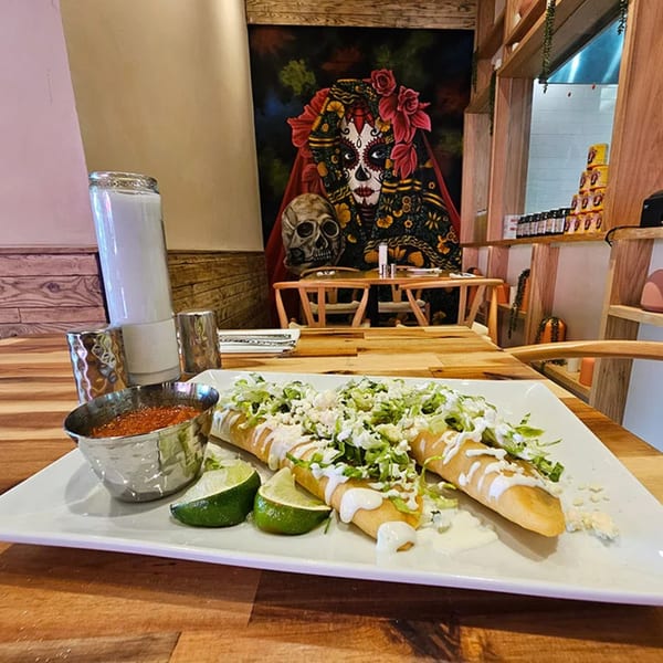 Okay Maguey's Mexican taquitos with lettuce, crema, and lime, with a Day of the Dead painting in the background.