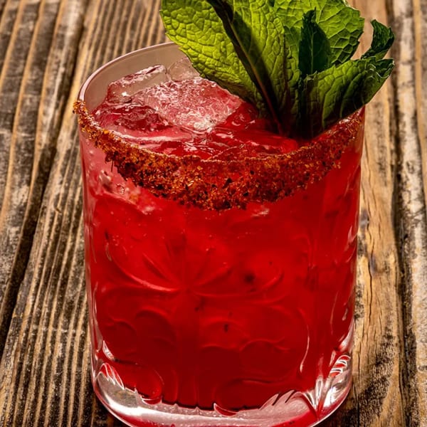 A vibrant red Mexican cocktail, garnished with fresh mint and a chili-salt rim, on a rustic wooden table.
