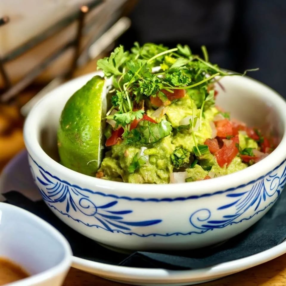 Okay Maguey's guacamole in a decorative bowl, garnished with cilantro, diced tomatoes, and a lime wedge.