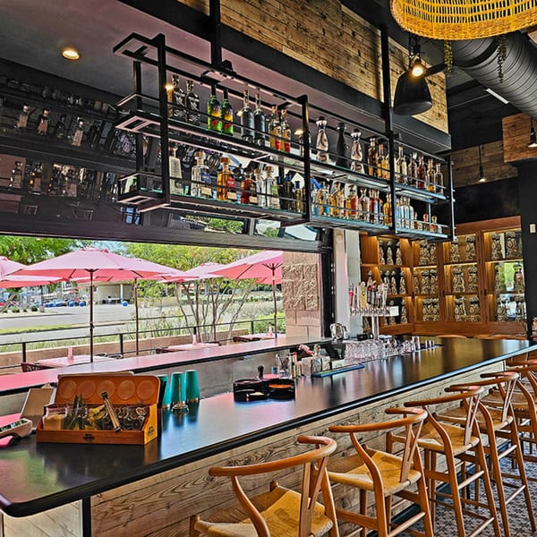 Okay Maguey's bar with liquor bottles, bar stools, and outdoor patio with pink umbrellas.