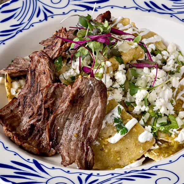 Grilled steak with chilaquiles, green sauce, crumbled cheese, and microgreens on a decorative plate.