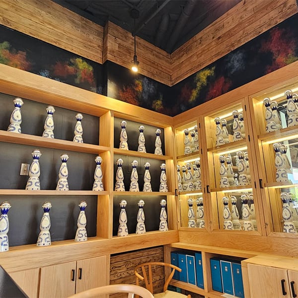 Okay Maguey's interior features wooden shelves displaying numerous decorative tequila bottles, with colorful wall art above.