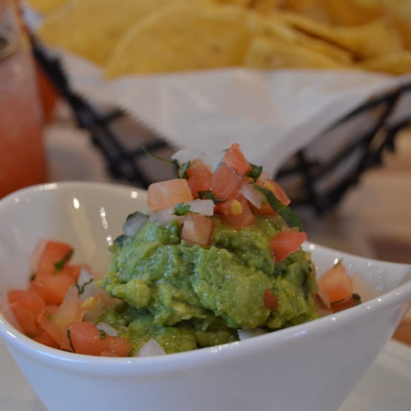 Okay Maguey's fresh guacamole topped with diced tomatoes, onions, and cilantro, with tortilla chips in the background.