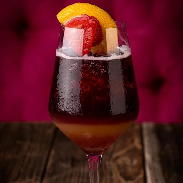 A two-toned cocktail with a dark red top and amber bottom, garnished with a lemon slice and strawberry.